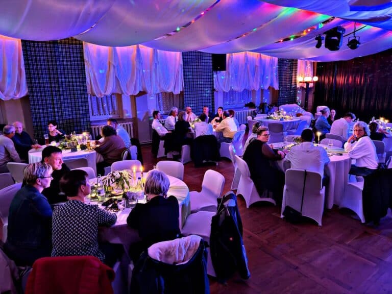 Eine Gruppe von Menschen sitzt um dekorierte Tische in einem warm beleuchteten Veranstaltungssaal mit drapierten Stoffen an der Decke und bunten Lichtern, die auf eine festliche Zusammenkunft oder Feier hindeuten