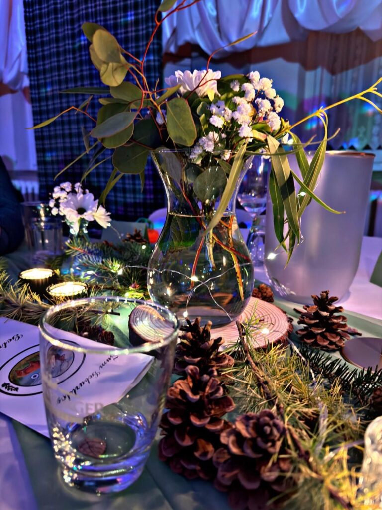 Eine Glasvase mit weißen Blumen und Grünzeug steht auf einem geschmückten Tisch mit Tannenzapfen, einem Glasbecher, immergrünen Zweigen, Kerzen und gefaltetem Papier und schafft eine festliche und gemütliche Atmosphäre