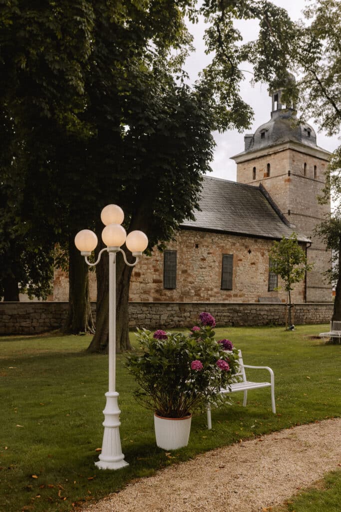 Eine weiße Bank und ein Laternenpfahl stehen auf einer Wiese neben einer blühenden Topfpflanze, mit einem steinernen Gebäude und hohen Bäumen im Hintergrund. Perfekter Ort für JGA Picknick.