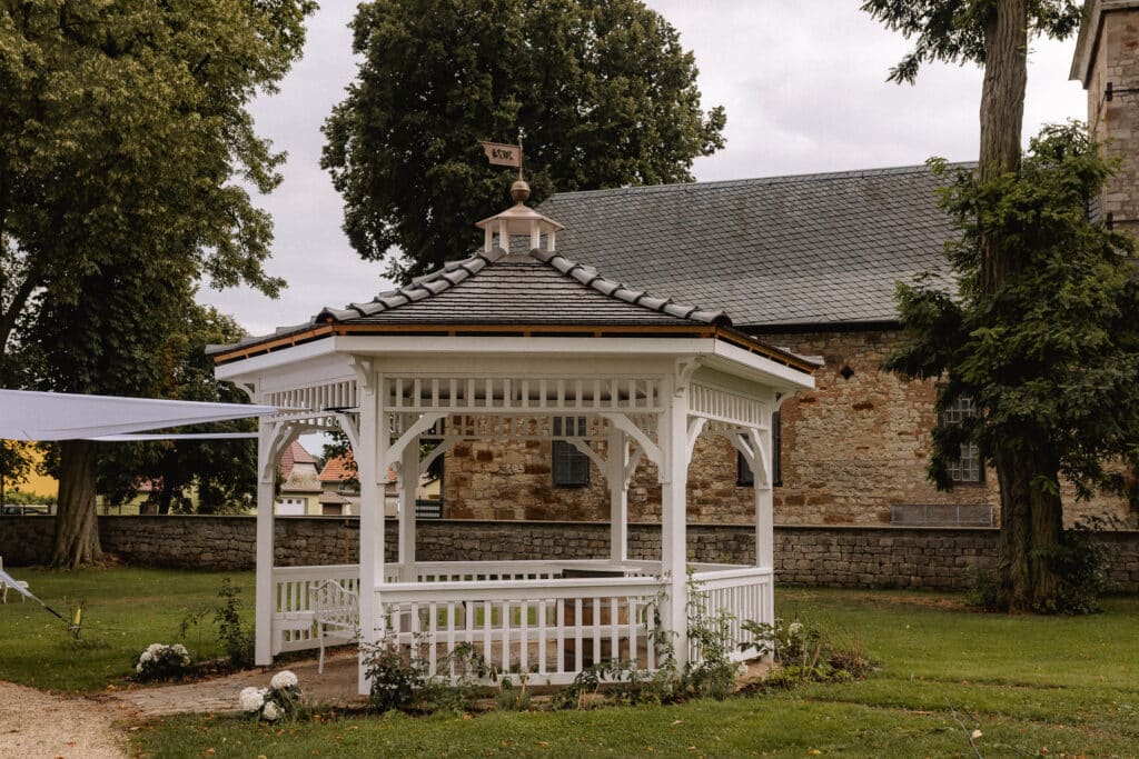 Ein weißer Holzpavillon mit einer Wetterfahne auf der Spitze steht an einem bewölkten Tag auf einer Wiese für JGA Picknick, umgeben von Bäumen, in der Nähe eines Steinhauses mit Ziegeldach.