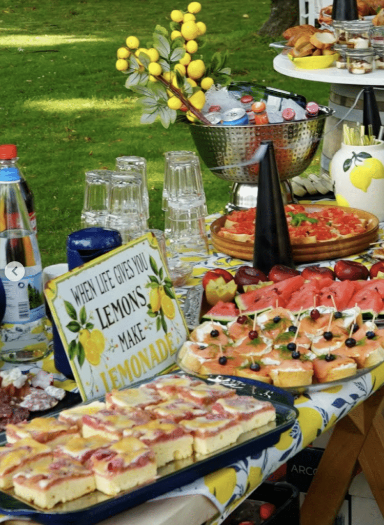 Auf einem farbenfrohen Tisch im Freien an einer Hochzeitslocation in Thüringen stehen Wassermelonenscheiben, Gebäck, Häppchen und Getränke in Flaschen bereit. Auf einem Schild steht: „Wenn dir das Leben Zitronen gibt, mach Limonade daraus.“ Außerdem sind Zitronen und Gläser zu sehen.