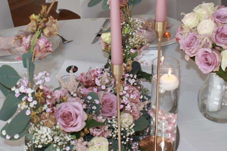 Eine Tischdekoration mit rosa Rosen, Schleierkraut, Grünpflanzen und zwei hohen rosa Kerzen in goldenen Kerzenhaltern. Diese elegante Dekoration eignet sich perfekt für eine Hochzeitslocation in Thüringen und besticht durch eine Schwimmkerze in einer Vase mit rosa Perlen, die für romantische Stimmung sorgt