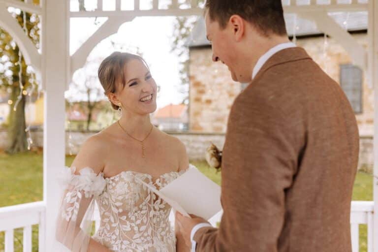 Eine Braut in einem Spitzenkleid lächelt den Bräutigam in einem braunen Anzug an, während dieser bei einer Hochzeitszeremonie im Freien unter einem weißen Pavillon aus einem Blatt vorliest