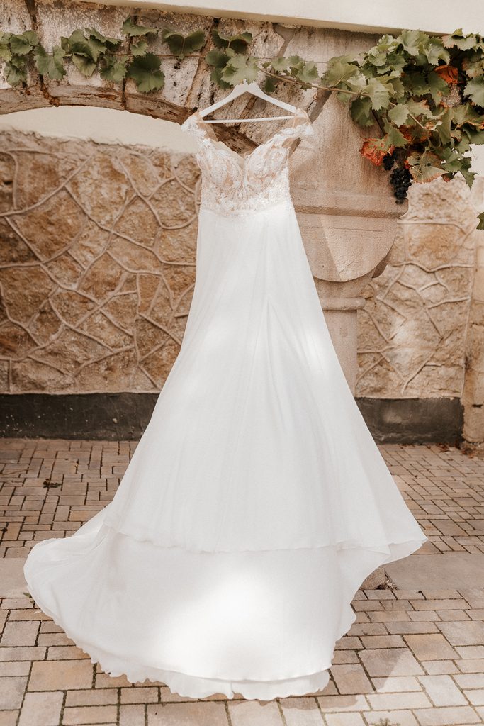 Ein weißes, wallendes Brautkleid mit Spitzendetails am Oberteil hängt draußen an einem Kleiderbügel an einer Steinmauer unter einem mit Weinreben bewachsenen Bogengang.