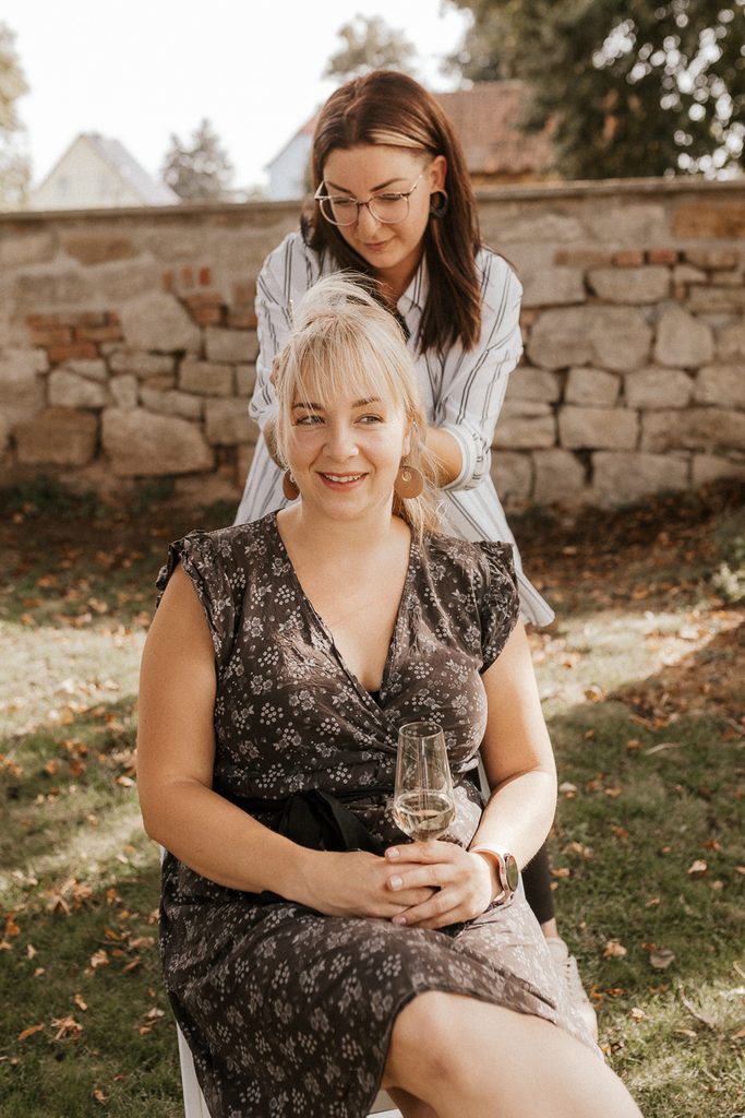 Eine Frau sitzt draußen und hält ein Glas Weißwein in der Hand, während eine andere Frau hinter ihr steht und ihr die Haare stylt. Sie befinden sich in einem sonnigen Garten mit einer Steinmauer und Bäumen im Hintergrund.