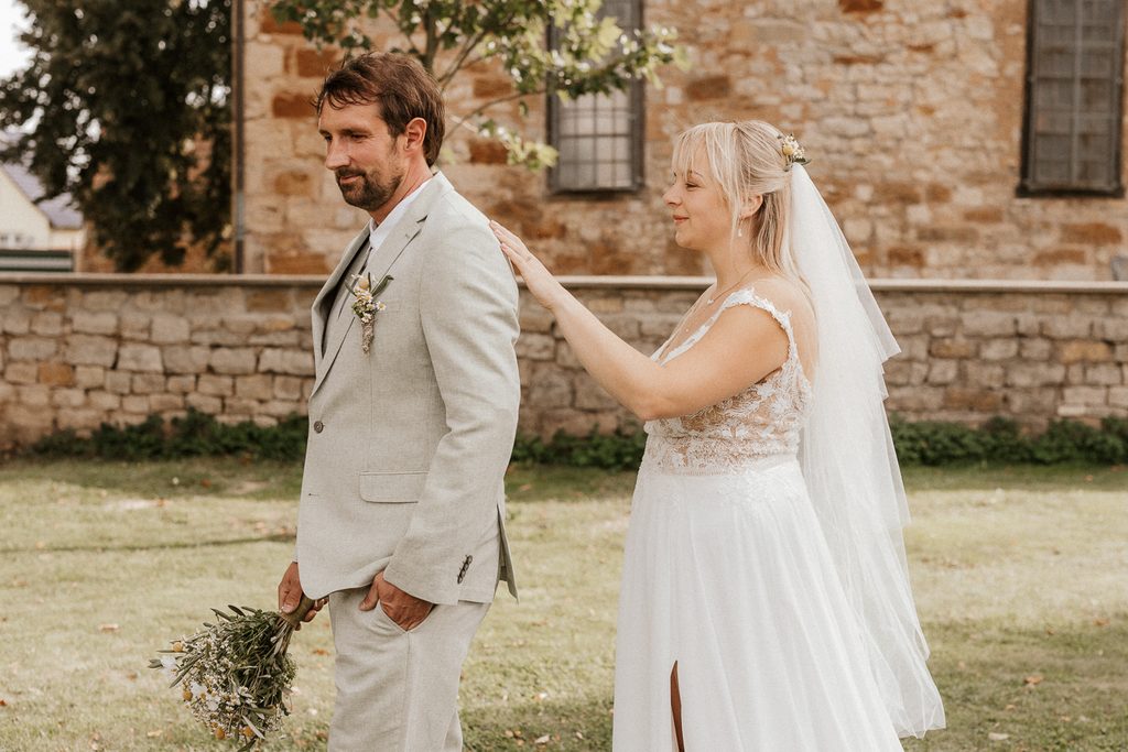 Eine Braut in einem weißen Kleid und mit Schleier berührt sanft die Schulter eines Bräutigams in einem hellen Anzug, der Blumen in der Hand hält; die Szene spielt im Freien in der Nähe einer Steinmauer und eines Gebäudes.