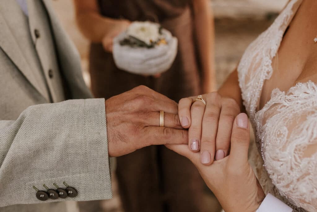 Nahaufnahme eines Brautpaares, das sich an den Händen hält und bei einer der vielen wunderschönen freien Trauungen seine Eheringe zeigt. Die Braut trägt ein Spitzenkleid, und im Hintergrund hält jemand ein Ringkissen in der Hand.