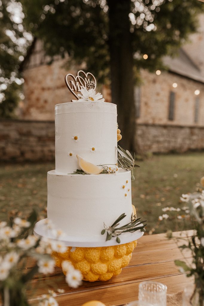 Eine zweistöckige weiße Hochzeitstorte mit Zitronen- und Gänseblümchenverzierungen steht im Freien auf einem gelben, strukturierten Ständer; darauf befindet sich ein Herzschild mit der Aufschrift „Mr & Mrs“, im Hintergrund sind verschwommene Bäume und ein Steingebäude zu sehen.