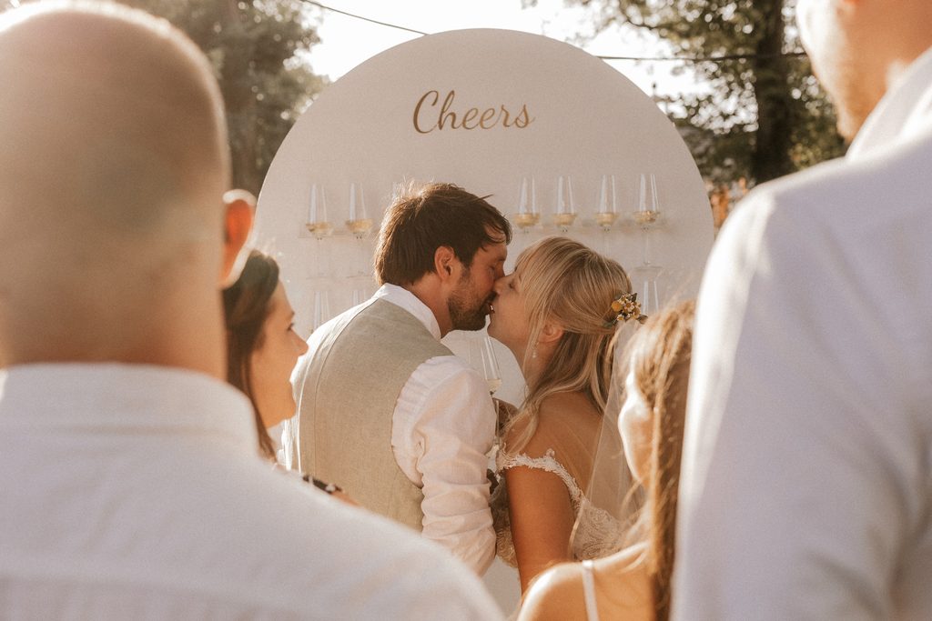 Ein Brautpaar küsst sich vor einem „Cheers“-Schild mit Champagnergläsern, umgeben von Gästen bei einer Hochzeit im Freien.