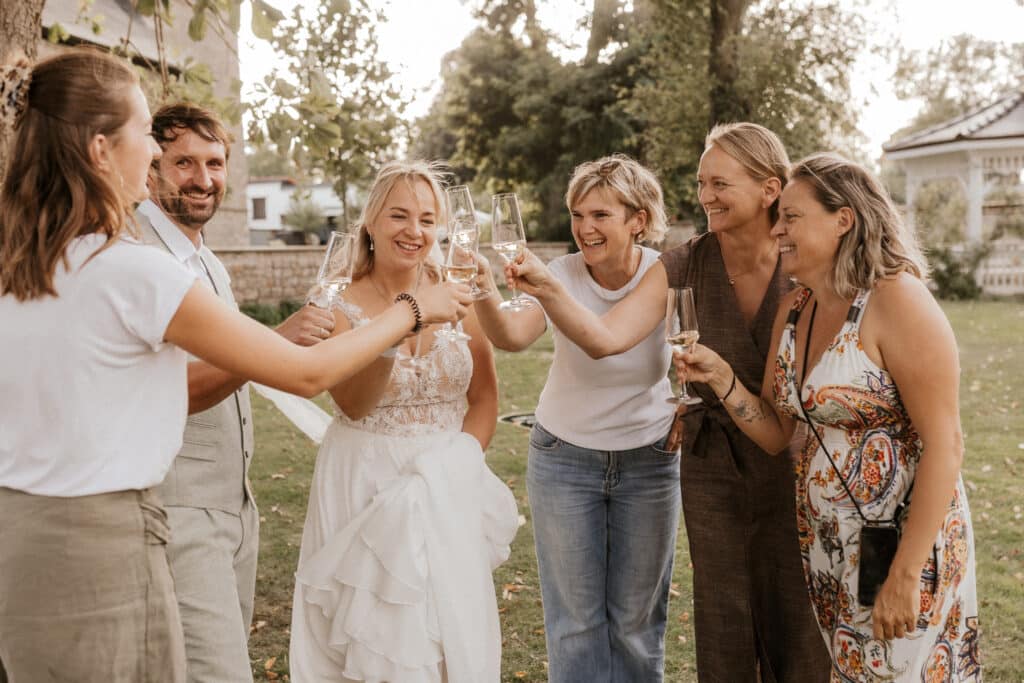 Eine Braut und fünf Gäste stehen im Freien, lächeln und stoßen mit Champagnergläsern an, um zu feiern. Im Hintergrund sind an einem sonnigen Tag Bäume und ein Pavillon zu sehen.