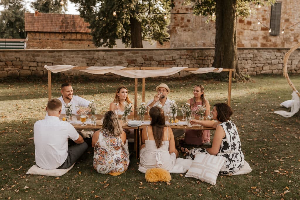 Sieben Personen sitzen auf Kissen um einen niedrigen Tisch im Freien herum, der mit Blumen und Getränken geschmückt ist, unter einem Holzvordach. Die Umgebung besteht aus einer Rasenfläche mit Bäumen, Backsteinmauern und Lichterketten, was den Eindruck einer entspannten, rustikalen Zusammenkunft vermittelt.