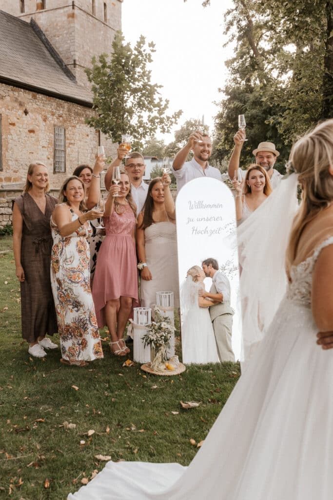 Eine Gruppe von Hochzeitsgästen in sommerlicher Kleidung erhebt im Freien bei einer der vielen „Freien Trauungen“ die Gläser zum Anstoßen, lächelt vor einem dekorativen Schild und Laternen, während im Vordergrund eine Braut in einem weißen Kleid steht.
