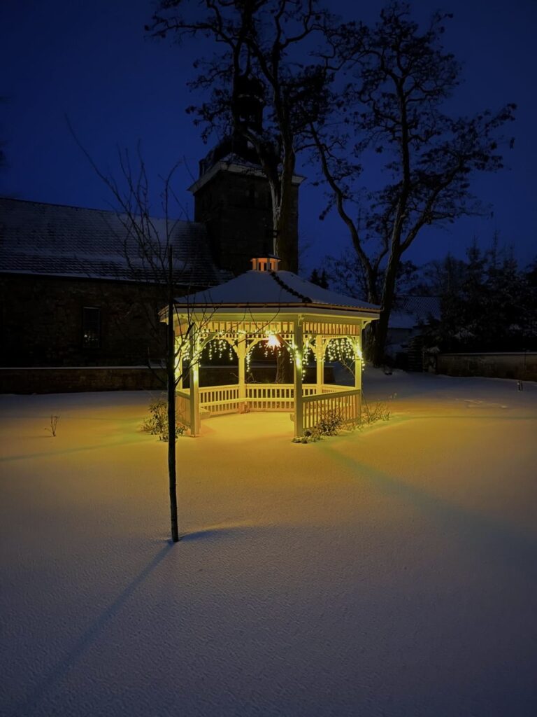 Ein nachts beleuchteter Pavillon in einem verschneiten Garten, im Hintergrund eine Steinkirche.