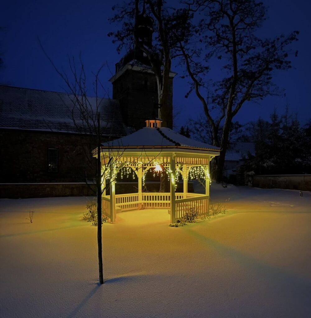 Ein nachts beleuchteter Pavillon in einem verschneiten Garten, im Hintergrund eine Steinkirche.