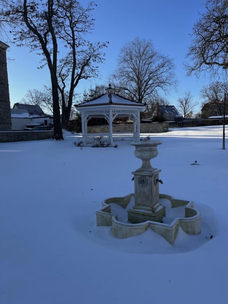 Im Vordergrund ein Steinbrunnen und im Hintergrund ein weißer Pavillon, beide umgeben von einer schneebedeckten Rasenfläche.