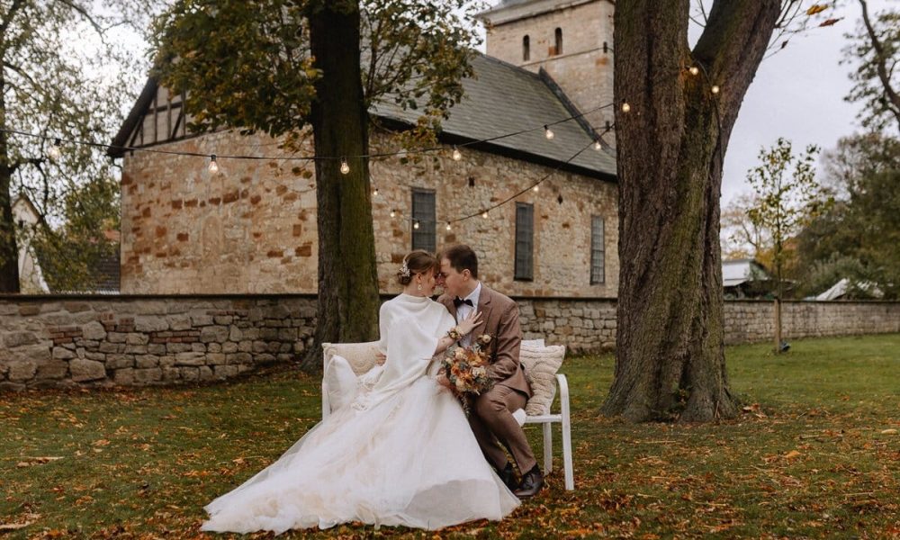 Eine Braut und ein Bräutigam sitzen eng beieinander auf einer Bank unter Lichterketten in einer grünen Umgebung, mit einer alten Steinkirche im Hintergrund. Die Braut hält einen Blumenstrauß in der Hand und trägt ein weißes Kleid; der Bräutigam trägt einen braunen Anzug.